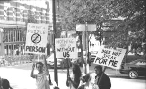 Description de l'image en noir et blanc : 3 manifestant(e)s autistes se tiennent debout sur le campus de l'université de Berkeley. Ils ont trois panneaux blancs de protestation où il est marqué en lettres majuscules "I'm not a puzzle, I am a person", "Nothing about us without us", "Autism Speaks does not Speak for me". Ils manifestent contre Autism Speaks.