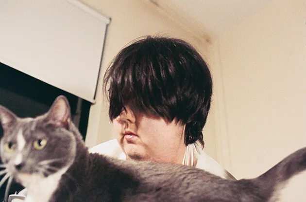 Photo de Mel baggs, iel a des cheveux noirs en coupe au bol qui lea couvrent les yeux. Iel se tient devant sa chatte grise et blanc.